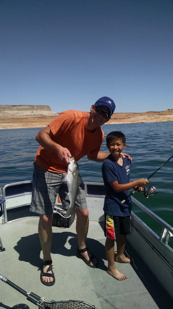 Image shows a young boy and a man holding a freshly caught fish on a sunny day, while fishing on the lake.